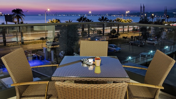 A balcony with wicker furniture, a table with condiments, and a view of the sea and city lights at dusk. - Hilton Garden Inn Izmir Bayrakli (Izmir)
