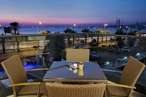 Un balcón con mobiliario de mimbre, una mesa con salsas y especias, y vistas al mar y las luces de la ciudad al anochecer.