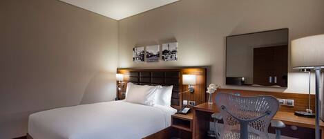 A hotel room with a large bed, bedside tables, a desk with a chair, a mirror, and wall-mounted artwork.