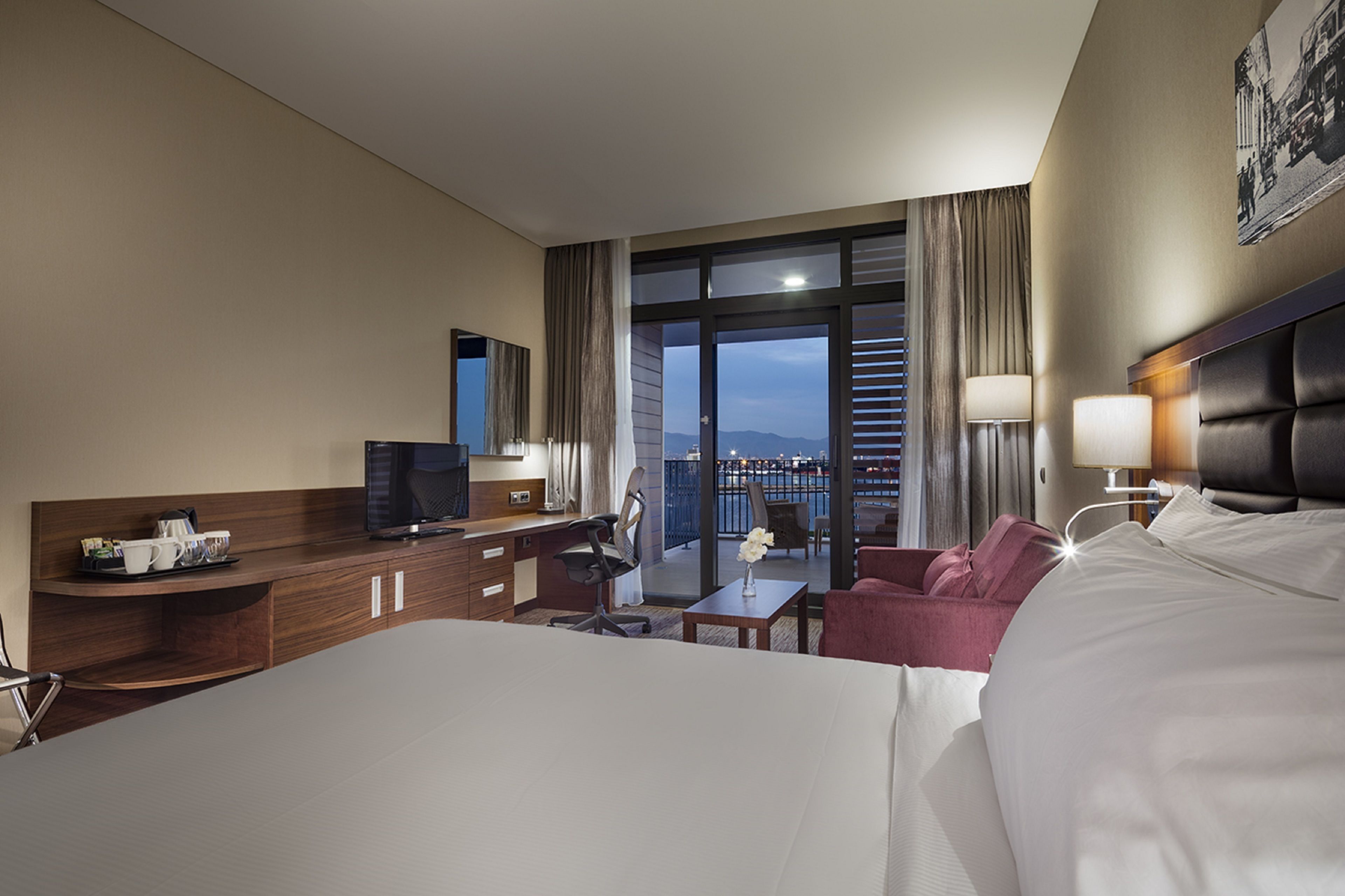 A hotel room with a large bed, a desk with a computer, a chair, a small table with a vase, and a view of the city through large windows.