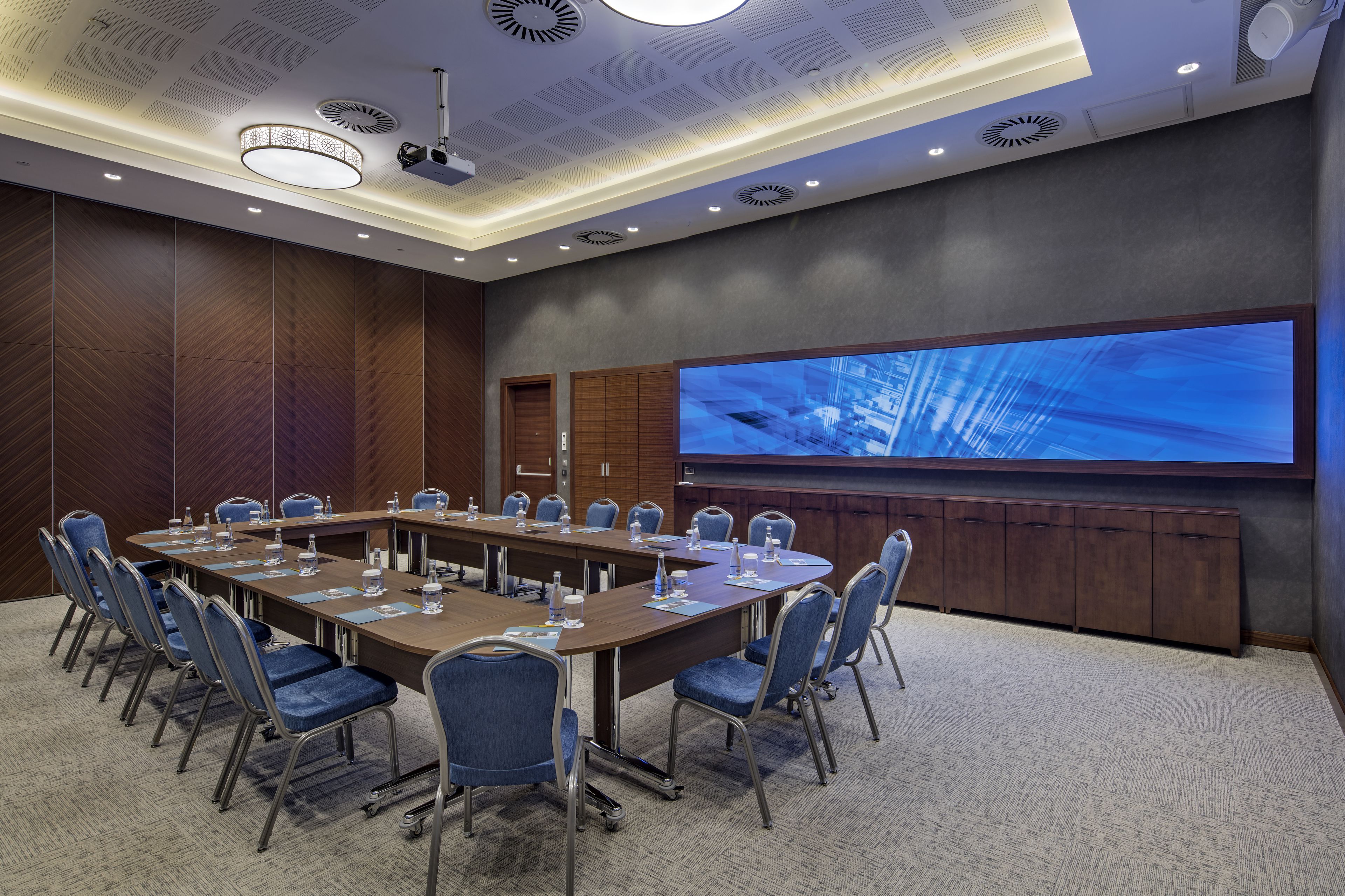 Uma sala de conferências com uma longa mesa, cadeiras, um ecrã grande e um teto com iluminação embutida e grelhas de ventilação do ar condicionado.