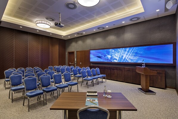 Una sala de conferencias con filas de sillas azules, un atril de madera y una pantalla grande que muestra una imagen.