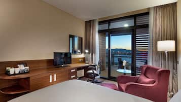 Habitación de hotel con cama, escritorio con computadora, una silla y vista de un paisaje urbano a través de grandes ventanas.