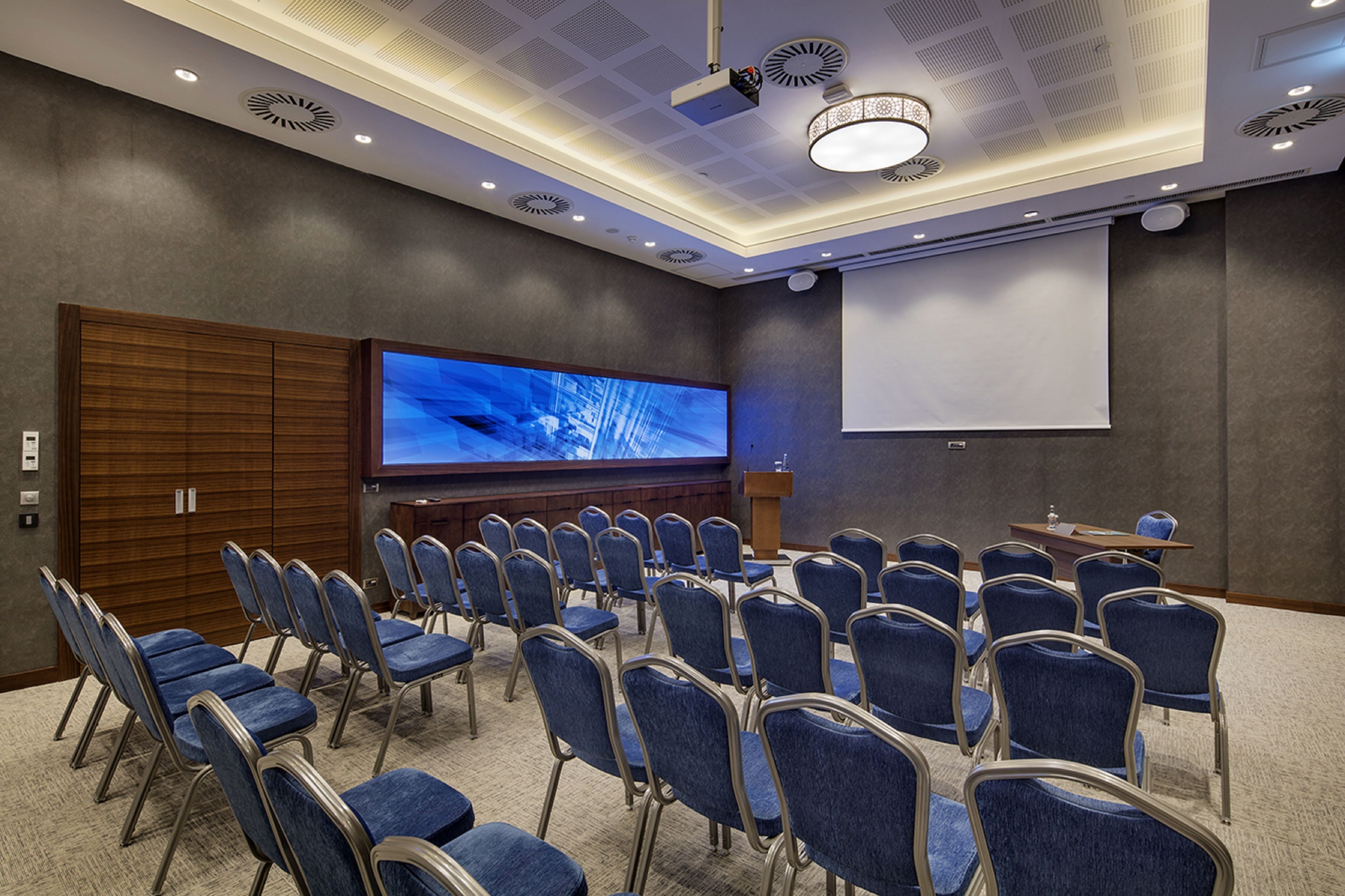 Une salle de conférence avec des rangées de chaises bleues, un grand écran et un projecteur.