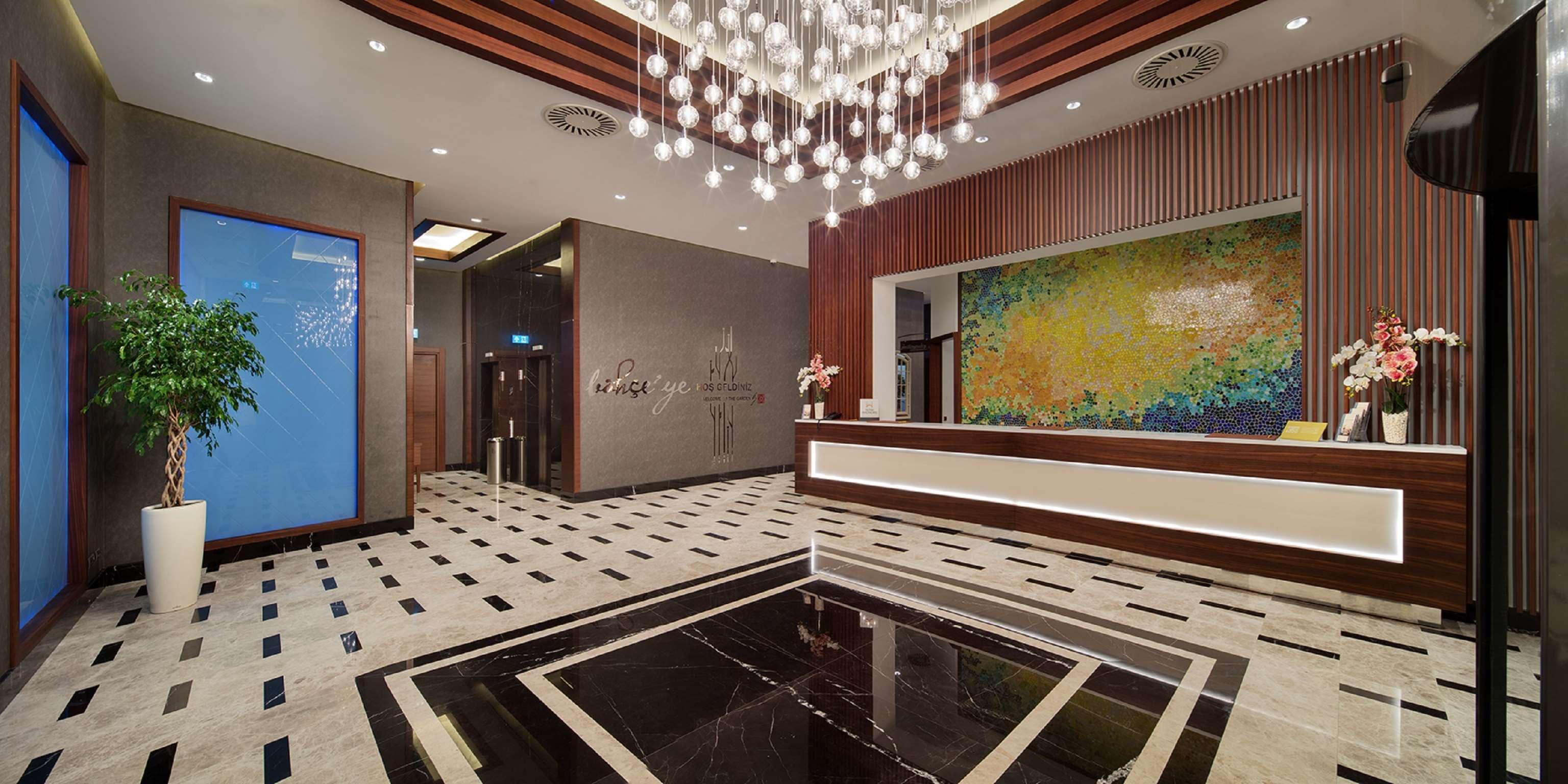 A modern hotel lobby with a reception desk, a large mosaic artwork, a potted plant, and a patterned floor.