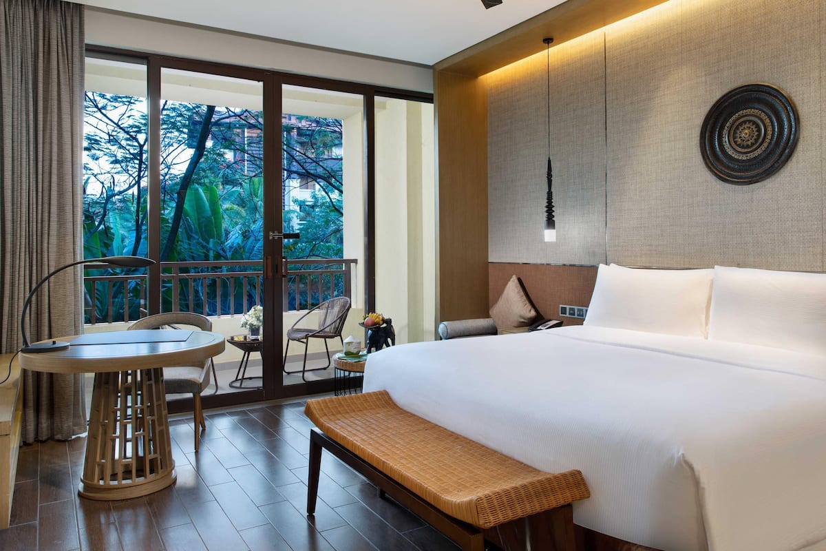 A hotel room with a bed, a round table, a bench, a ceiling fan, and a view of greenery outside.