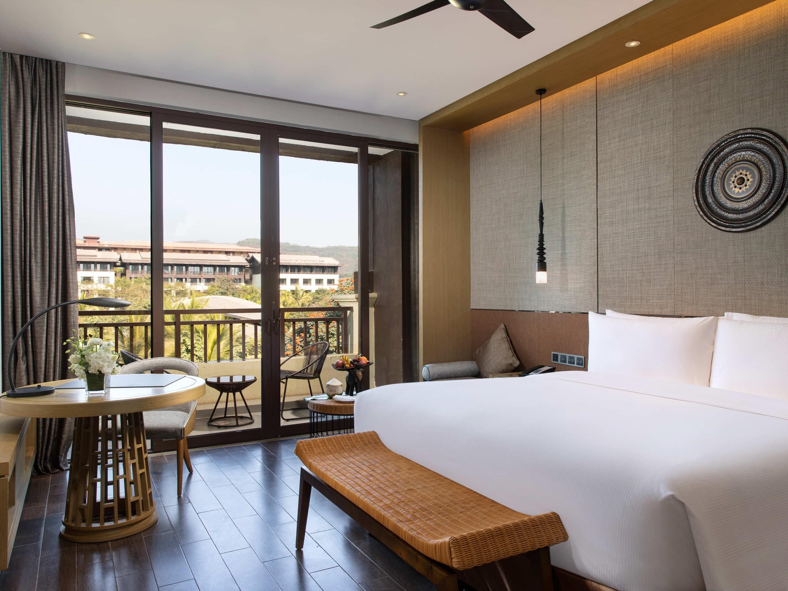 A hotel room with a large bed, a small table with a vase of flowers, a bench, and a balcony with a view of buildings and trees.