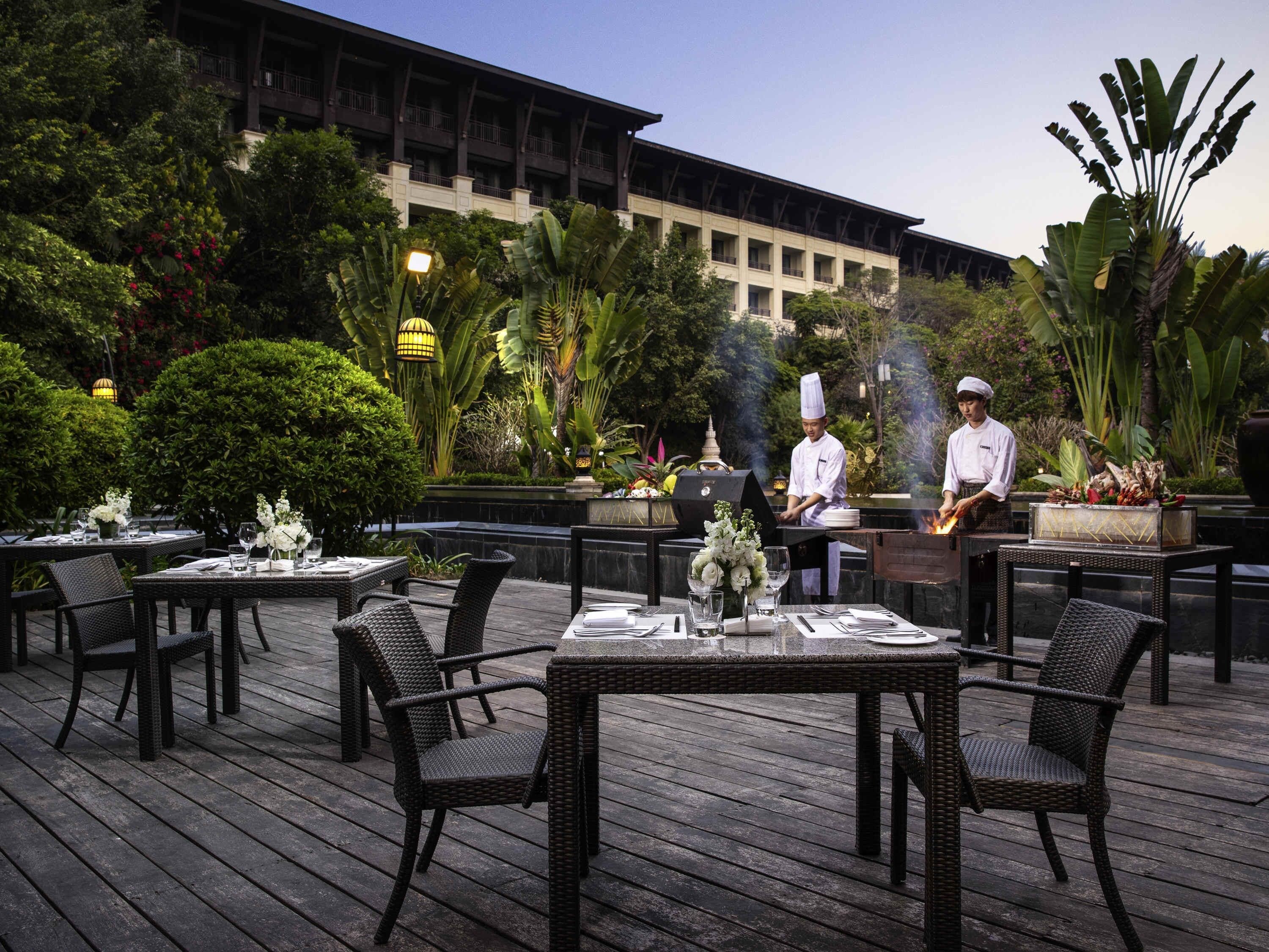 An outdoor dining area with wicker chairs and tables, chefs preparing food, and lush greenery in the background.