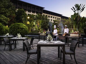 Un área de comedor al aire libre con sillas y mesas de mimbre, chefs preparando comida y exuberante follaje de fondo.