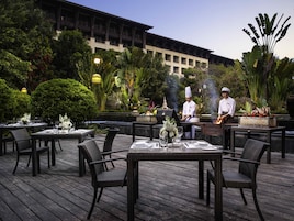An outdoor dining area with wicker chairs and tables, chefs preparing food, and lush greenery in the background.