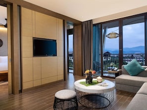 A modern hotel room with a flat-screen TV, a round coffee table with a decorative elephant figurine, a sofa with a green pillow, and a balcony with a view of the mountains.