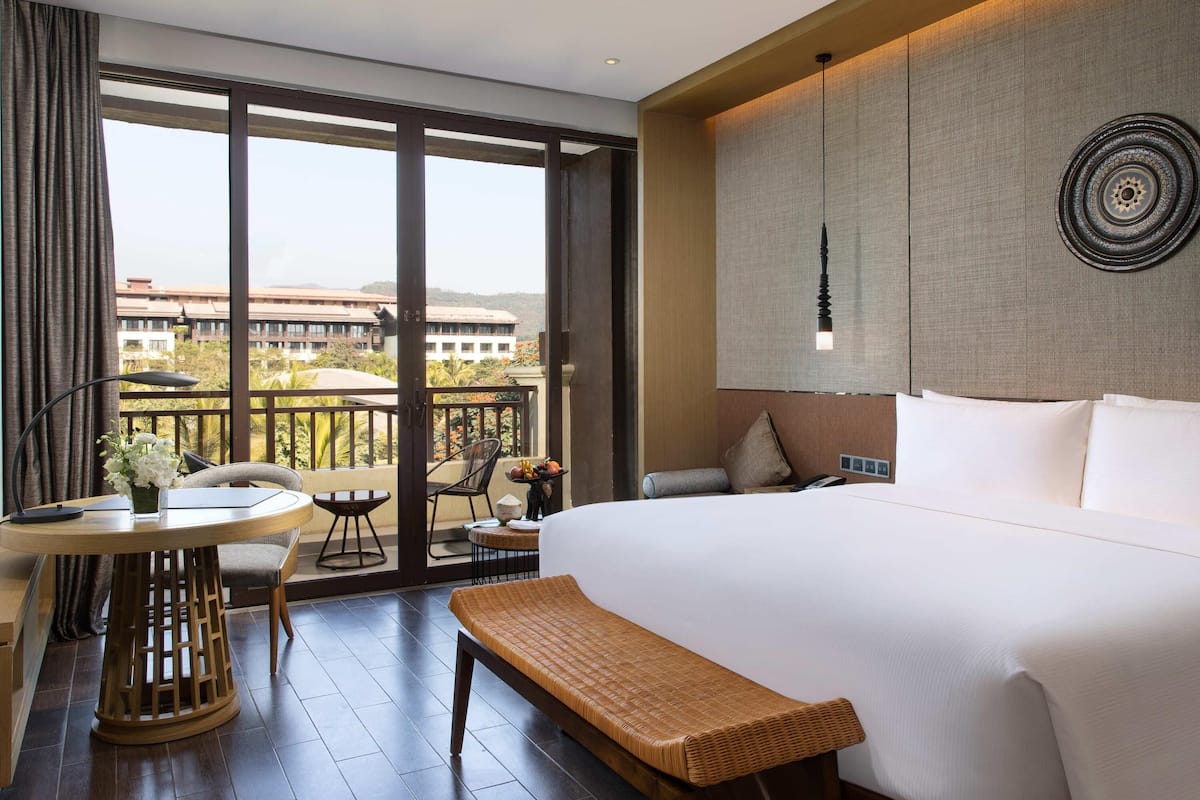 A hotel room with a large bed, a small table with a vase of flowers, a bench, and a balcony with a view of buildings and trees.