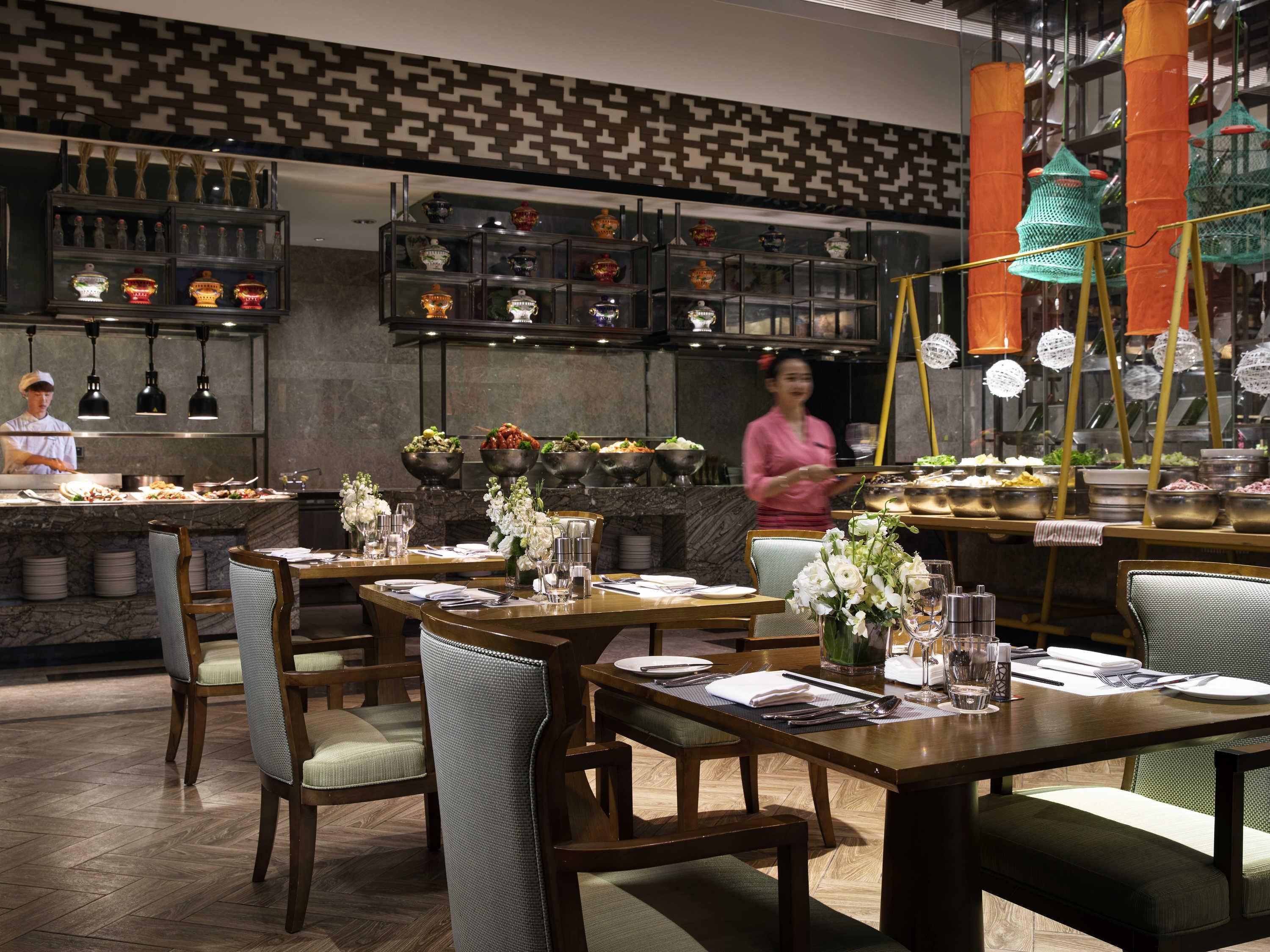 A restaurant interior with wooden tables, chairs, and a bar area with shelves of bottles and decorative items.