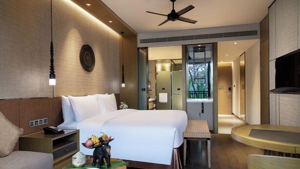 A modern hotel room with a large bed, bedside table, and a small round table. There is a ceiling fan, a wall-mounted light, and a view of the bathroom through an open door. - Pullman Resort Xishuangbanna (Xishuangbanna Dai)