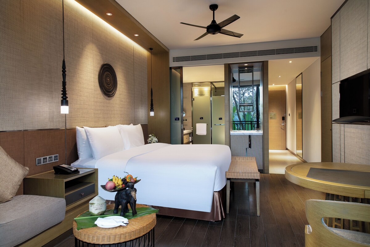 A modern hotel room with a large bed, bedside table, and a small round table. There is a ceiling fan, a wall-mounted light, and a view of the bathroom through an open door.