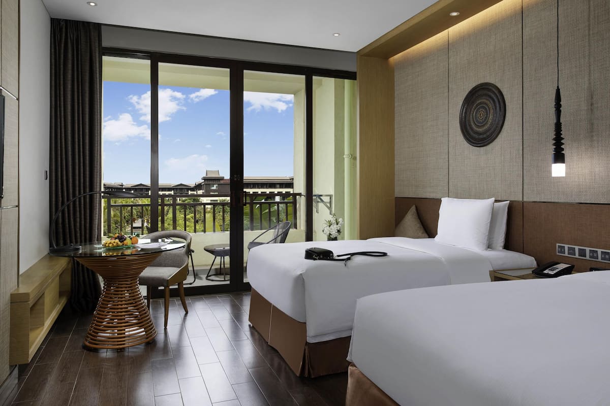 A hotel room with two beds, a dining table with fruit, a balcony with a view, and a ceiling fan.