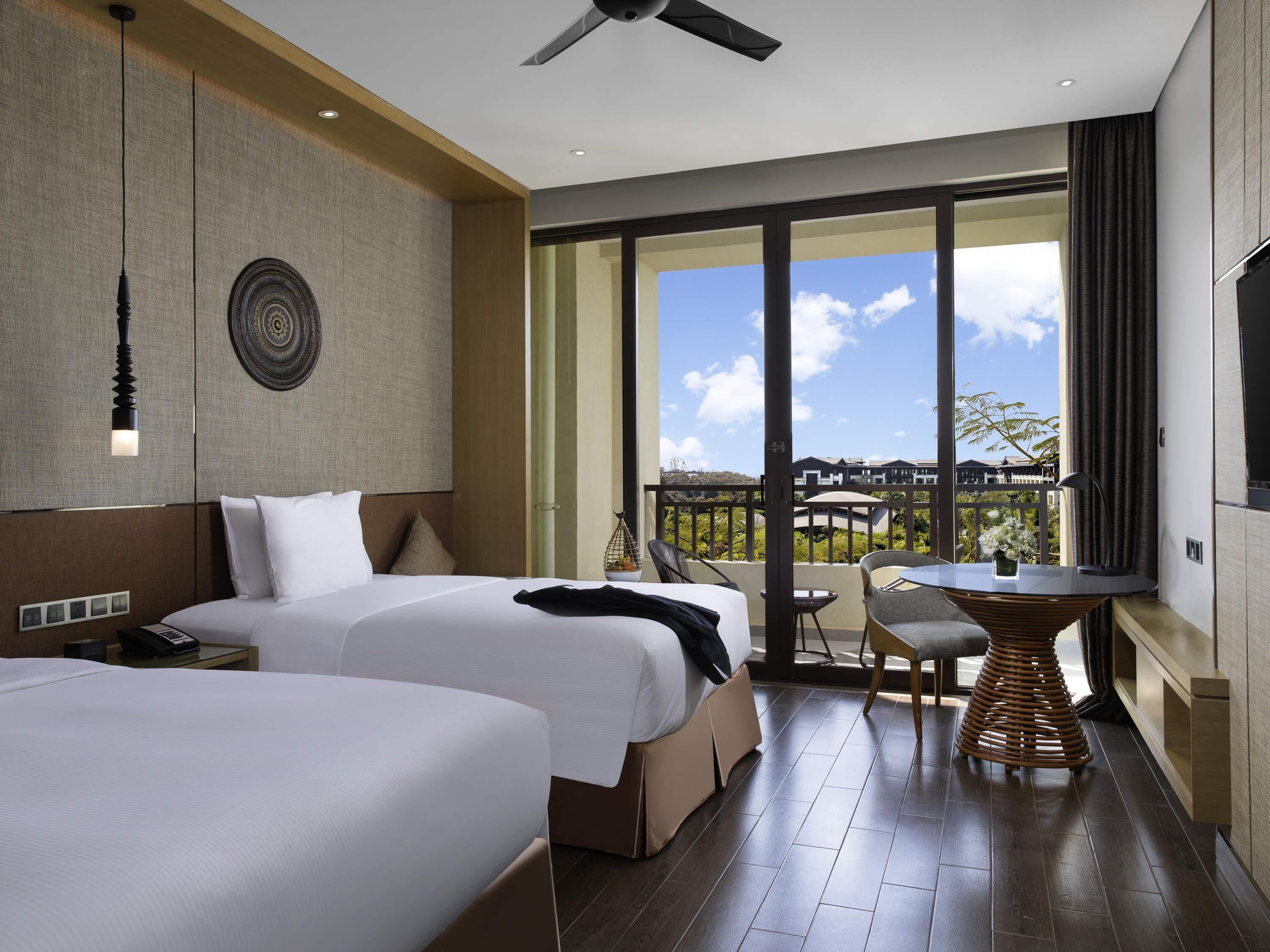 A hotel room with two beds, a balcony with a table and chairs, a ceiling fan, and a large window offering a view.