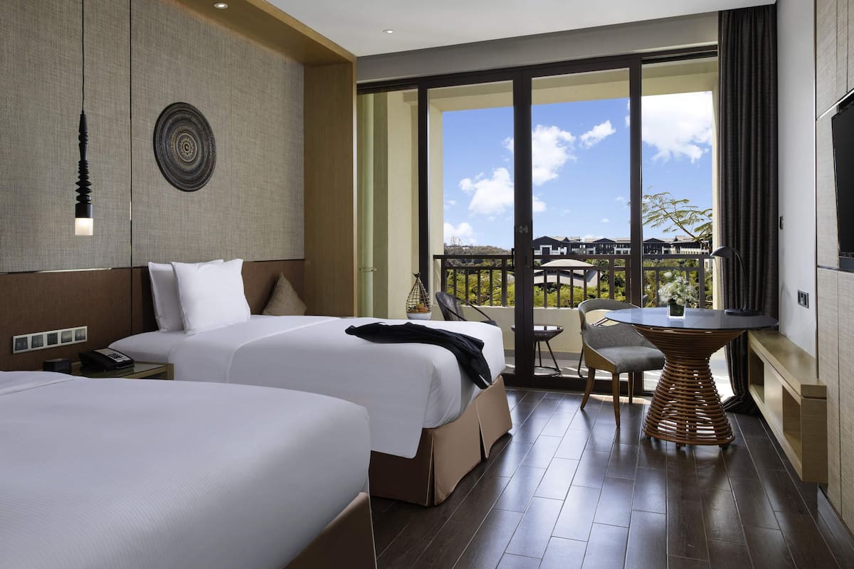 A hotel room with two beds, a balcony with a table and chairs, a ceiling fan, and a large window offering a view.