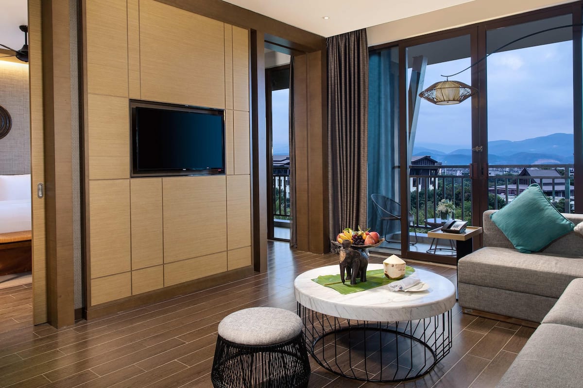 A modern hotel room with a flat-screen TV, a round coffee table with a fruit centerpiece, a sofa with a green pillow, and a balcony with a view of mountains.