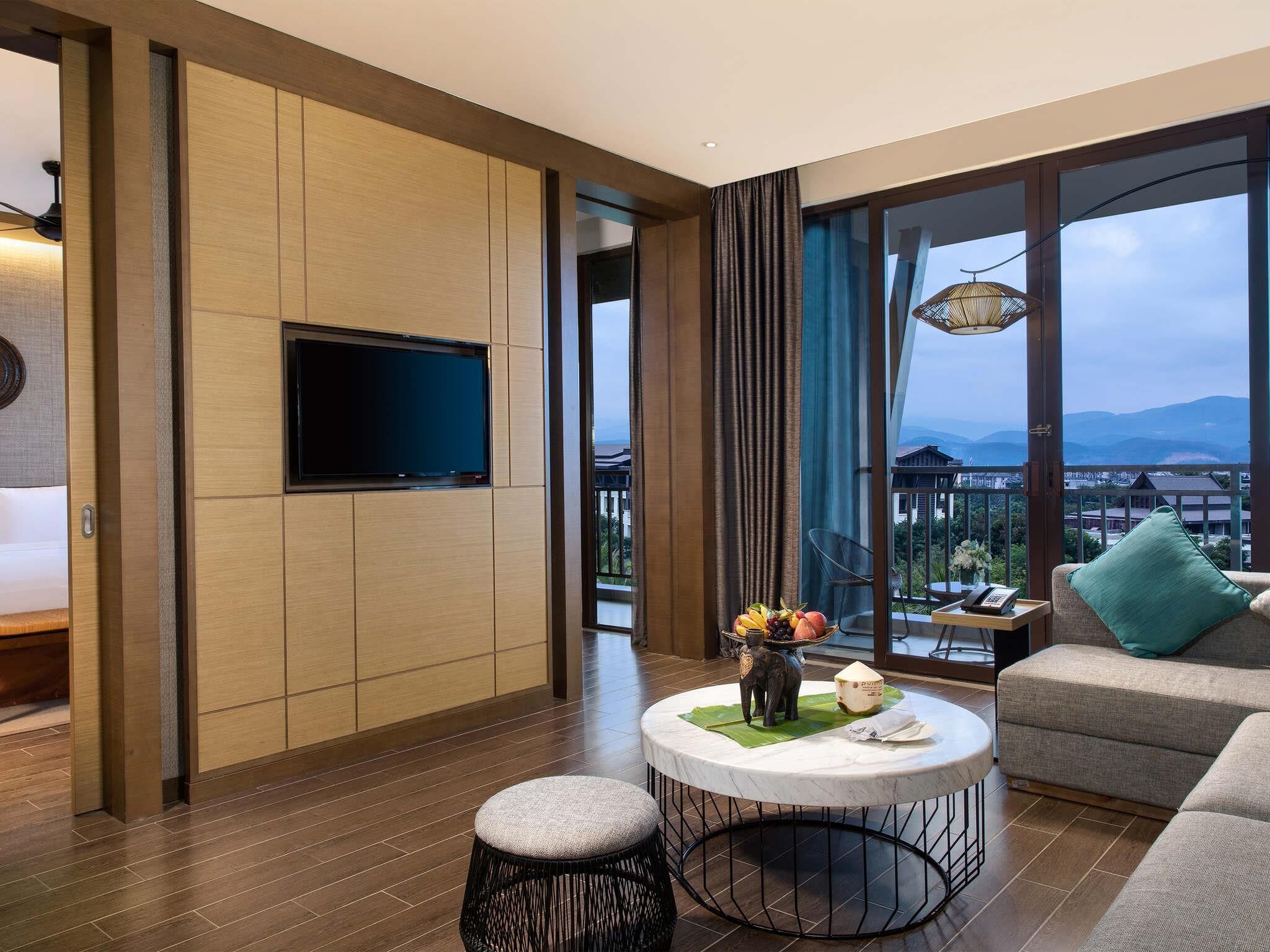 A modern hotel room with a flat-screen TV, a round coffee table with a decorative elephant figurine, a sofa with a green pillow, and a balcony with a view of the mountains.