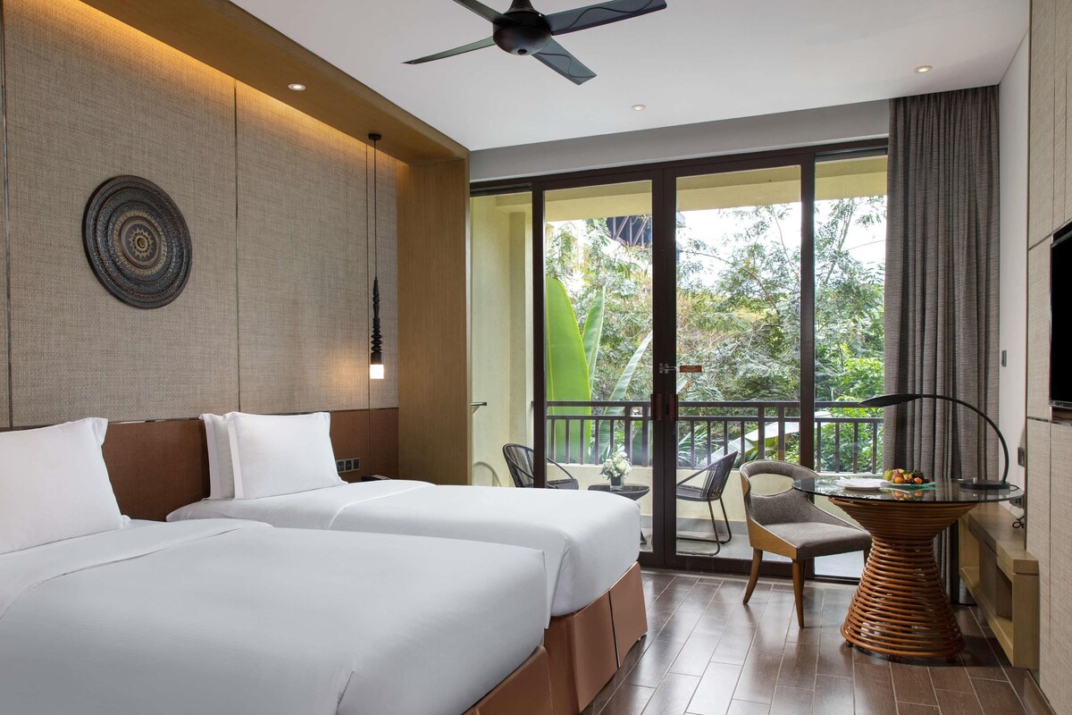 A hotel room with two beds, a balcony with a table and chairs, and a view of greenery outside.