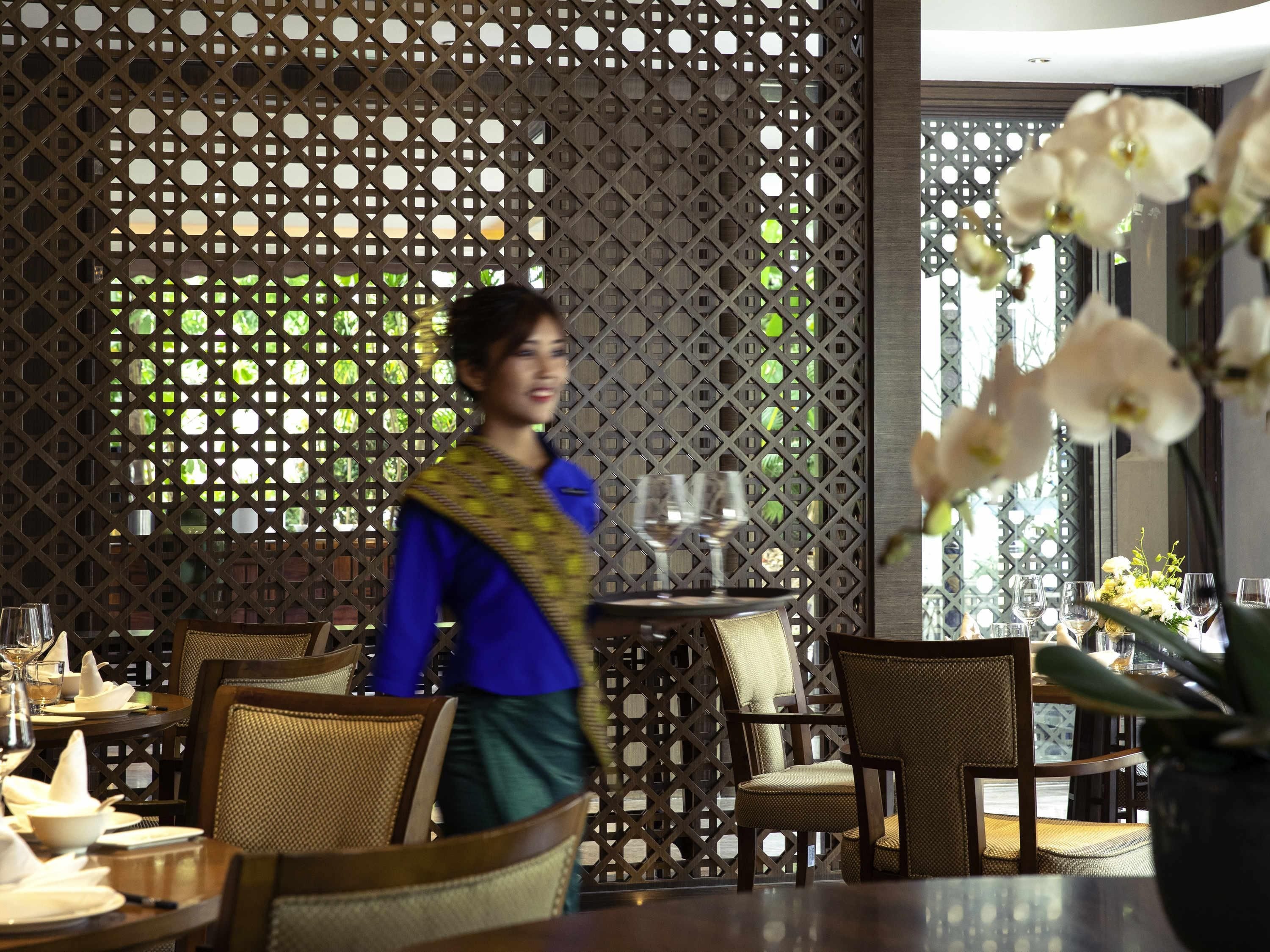 A restaurant interior with a lattice partition, dining tables with chairs, and a person in traditional attire.