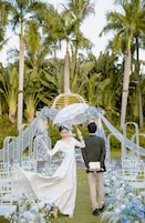 Una ceremonia de boda con una novia y un novio bajo un parasol blanco, rodeados de sillas blancas y decoraciones florales.