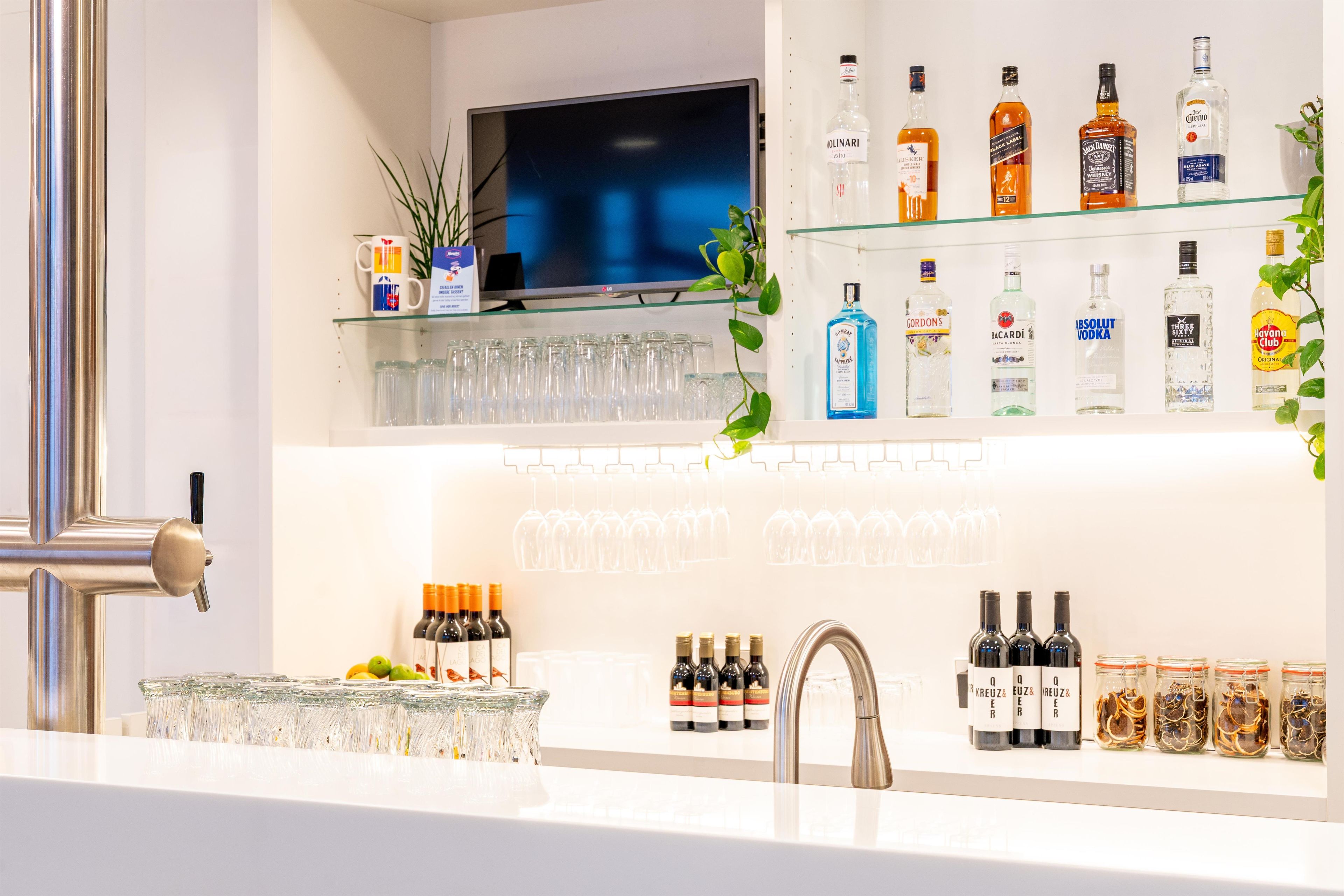 Uma área de bar com prateleiras cheias de garrafas de bebidas, copos e uma televisão.