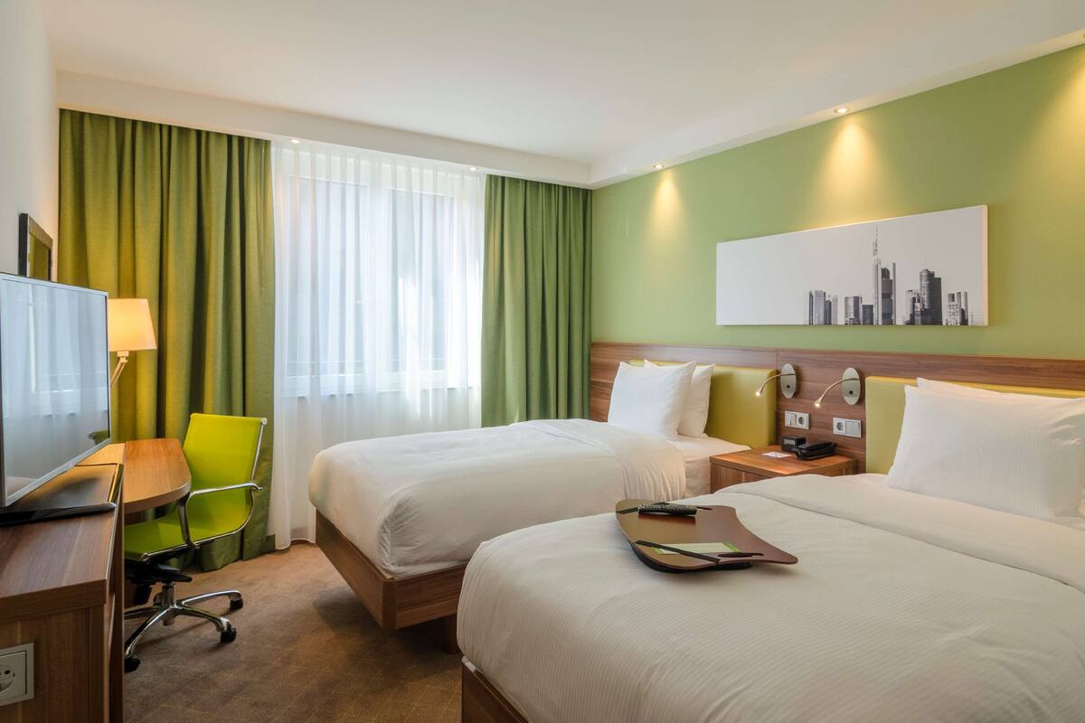 A hotel room with two beds, a desk with a chair, a television, and a cityscape painting on the wall.