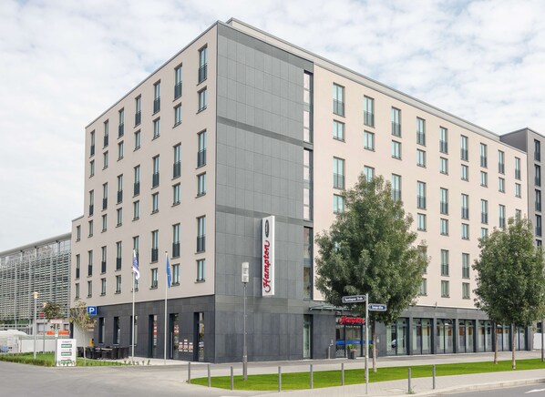 A multi-story hotel with a modern design, featuring a prominent entrance with a sign, a parking area, and a well-maintained lawn. - Hampton by Hilton Frankfurt City Centre (Frankfurt)