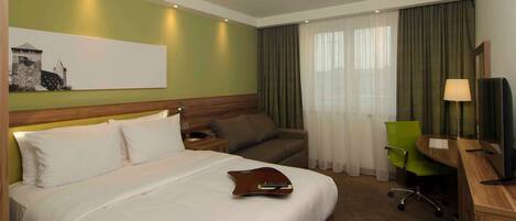 A hotel room with a bed, a desk, a chair, a television, and a painting on the wall.