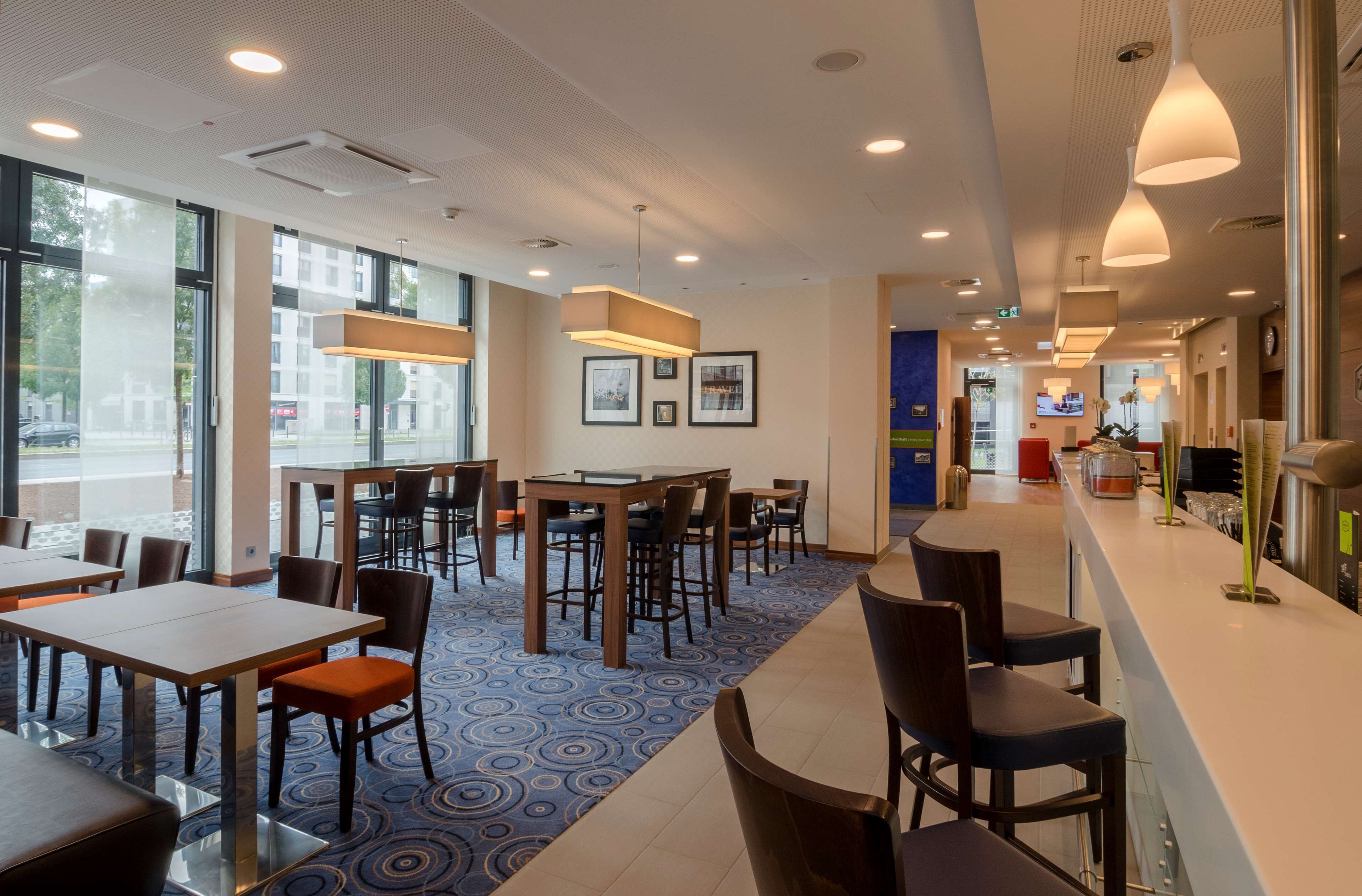 A modern hotel lobby with tables and chairs, large windows, and a patterned carpet.