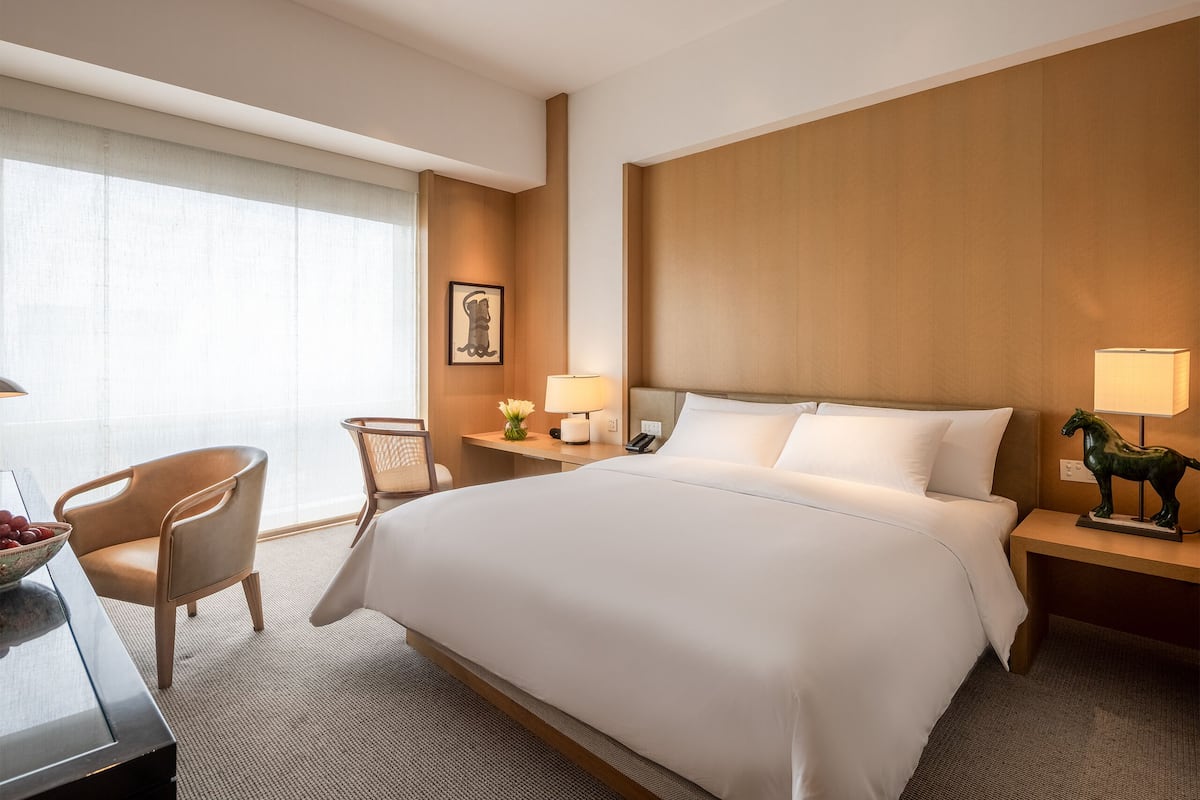A hotel room with a large bed, a desk with a lamp, a chair, and a small table with a lamp.