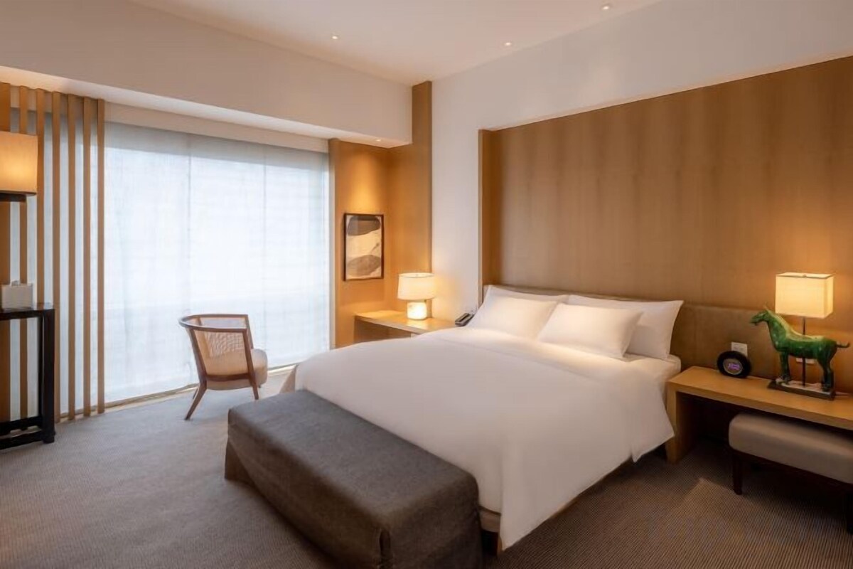Ein Hotelzimmer mit einem großen Bett, Nachttischen, einem Sessel und einem Fenster mit Vorhängen.