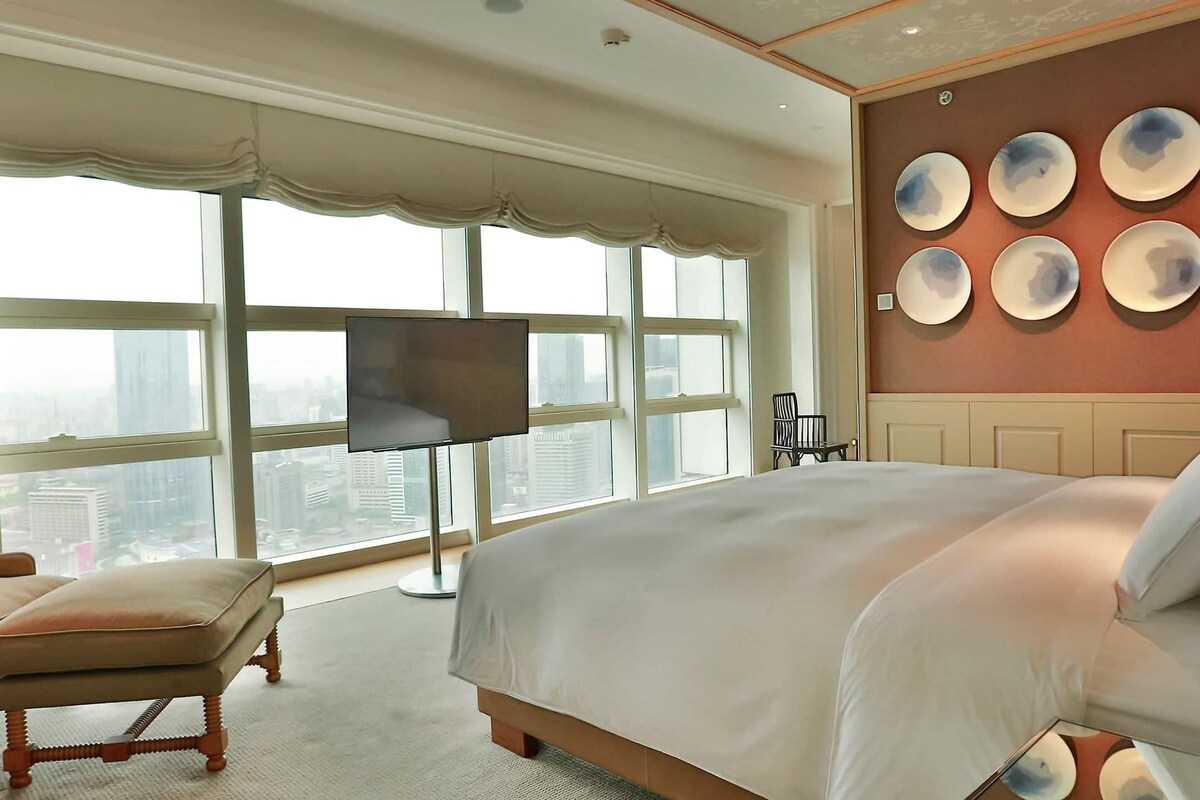 Ein geräumiges Schlafzimmer mit einem großen Bett, Blick auf die Stadt durch raumhohe Fenster und einer Wand, die mit runden Tellern dekoriert ist.