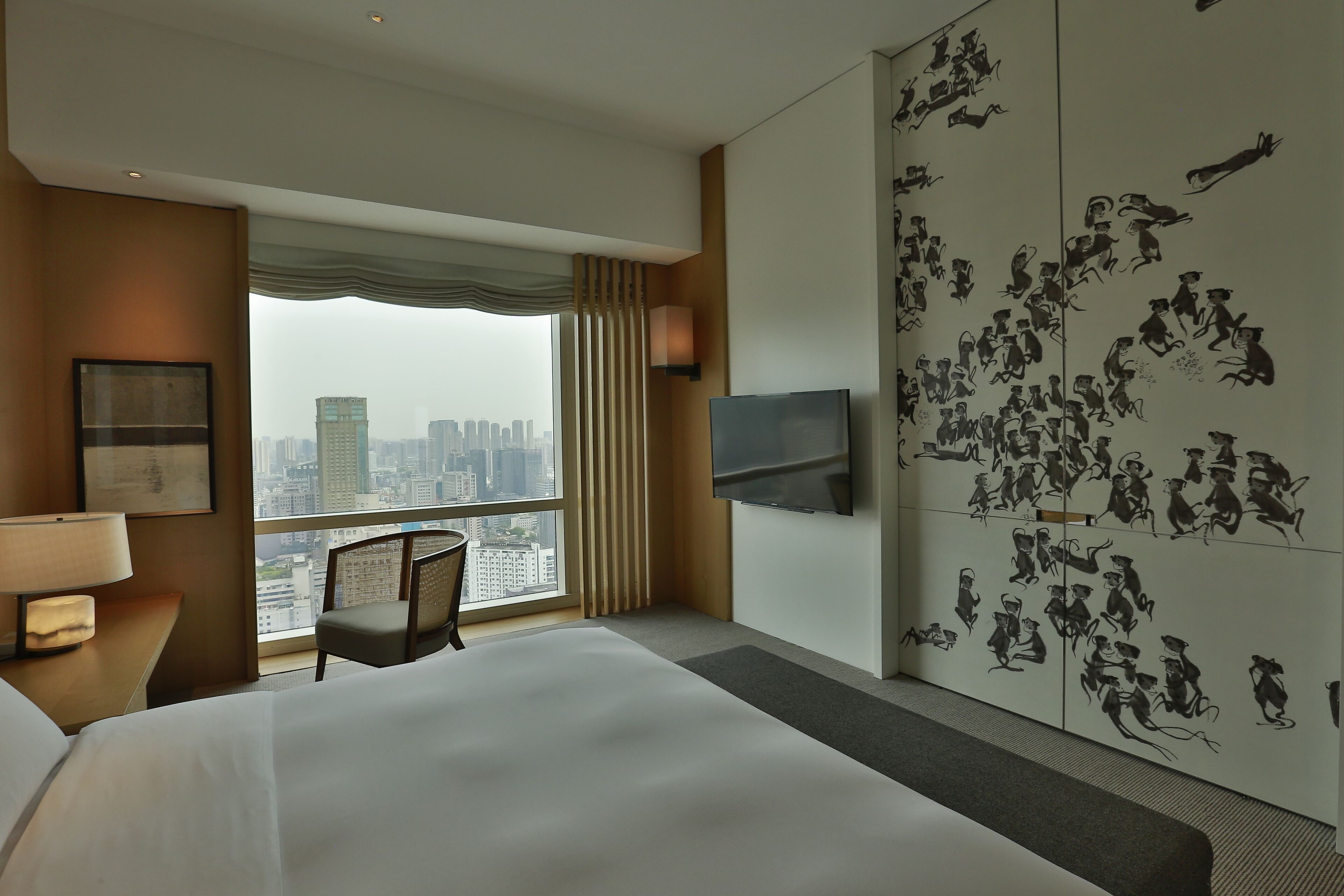 Une chambre d’hôtel avec un grand lit, une chaise, un bureau avec une lampe, un téléviseur fixé au mur et une vue sur la ville par la fenêtre.