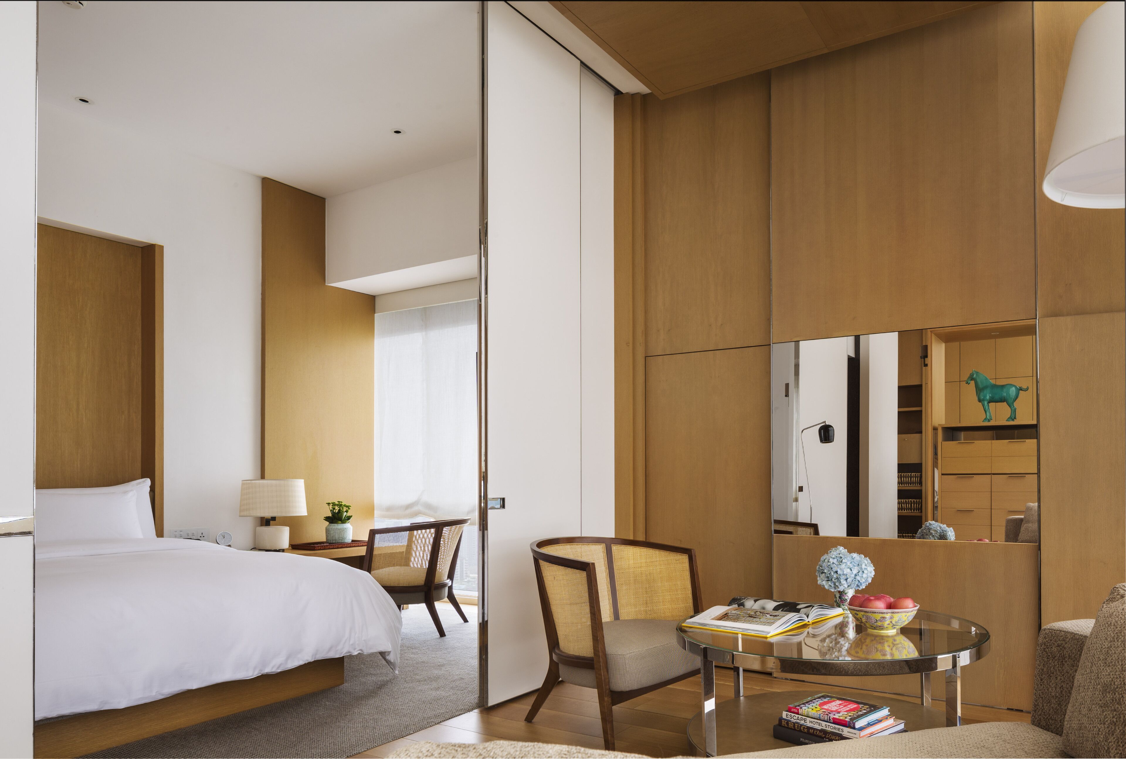 A modern hotel room with a bed, a desk with a lamp, a chair, a small table with books, and a decorative item.