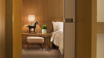A hotel room with a wooden headboard, a bedside lamp, a small table with a horse figurine and a plant, a white bed, and a bench.