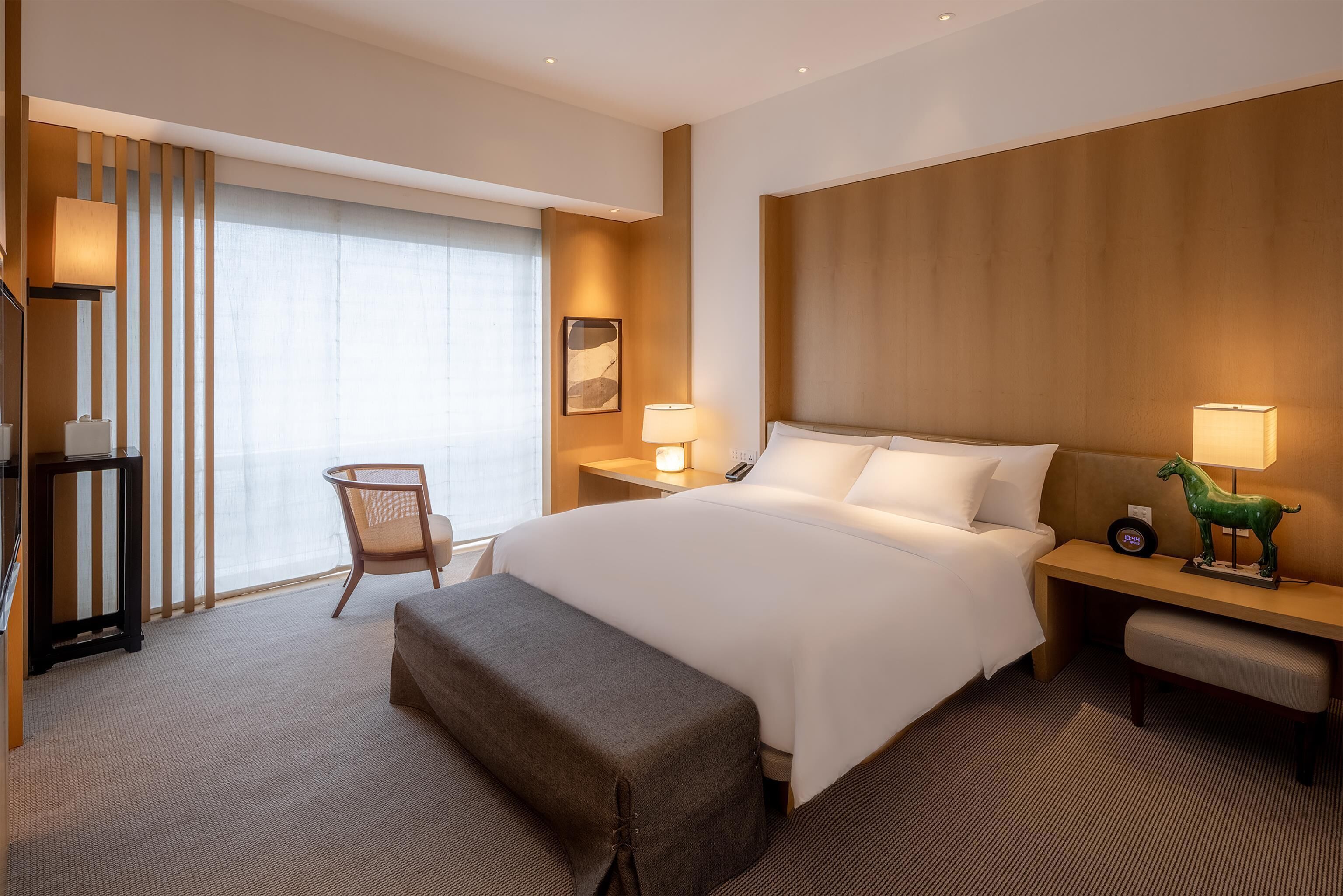A hotel room with a large bed, a chair, a desk, a lamp, and a window with blinds.