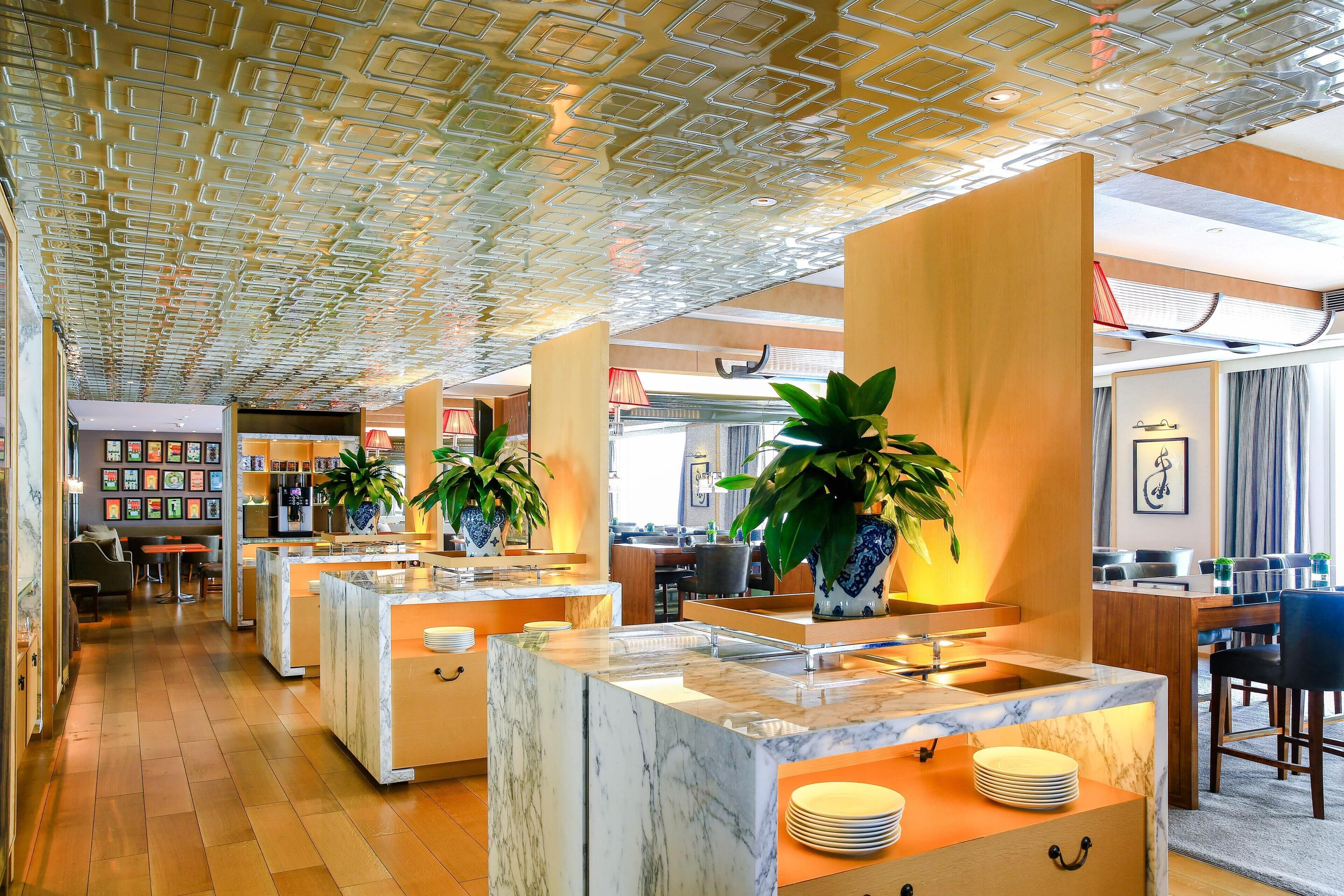 A modern hotel lobby with marble counters, potted plants, and a patterned ceiling.