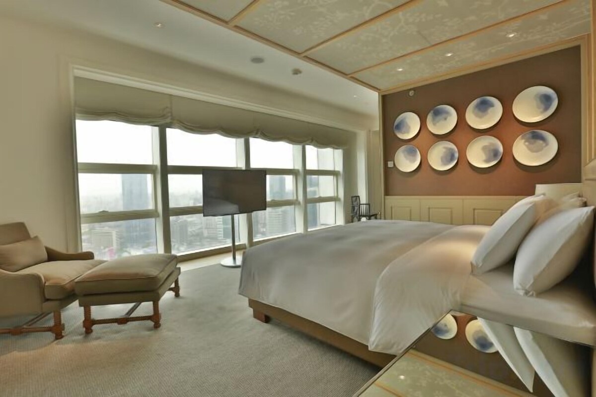 Ein geräumiges Schlafzimmer mit einem großen Bett, einer Sitzecke mit Sessel und Hocker, einem Flachbildfernseher und Blick auf die Stadt durch große Fenster.
