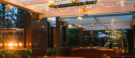 Un área de estar al aire libre con bancos y mesas de madera, luces colgantes y techo de vidrio.