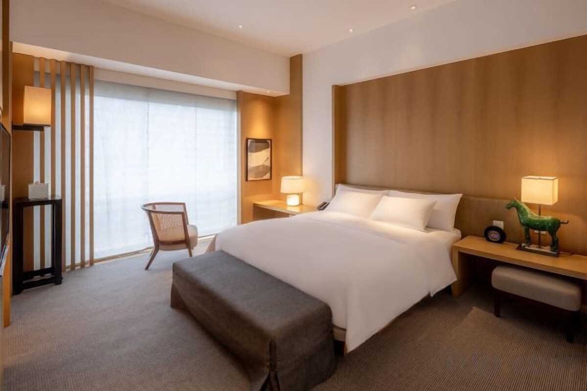 Ein Hotelzimmer mit einem großen Bett, Nachttischen, einem Sessel und einem Fenster mit Vorhängen.