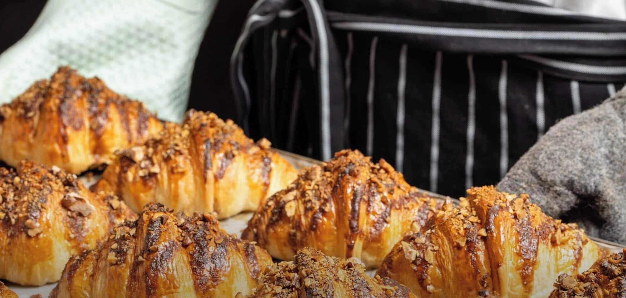 A tray of croissants with a golden-brown crust, likely freshly baked, with a sprinkling of nuts on top.