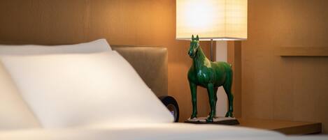 A bed with white linens, a green horse-shaped lamp, and a wall-mounted clock.