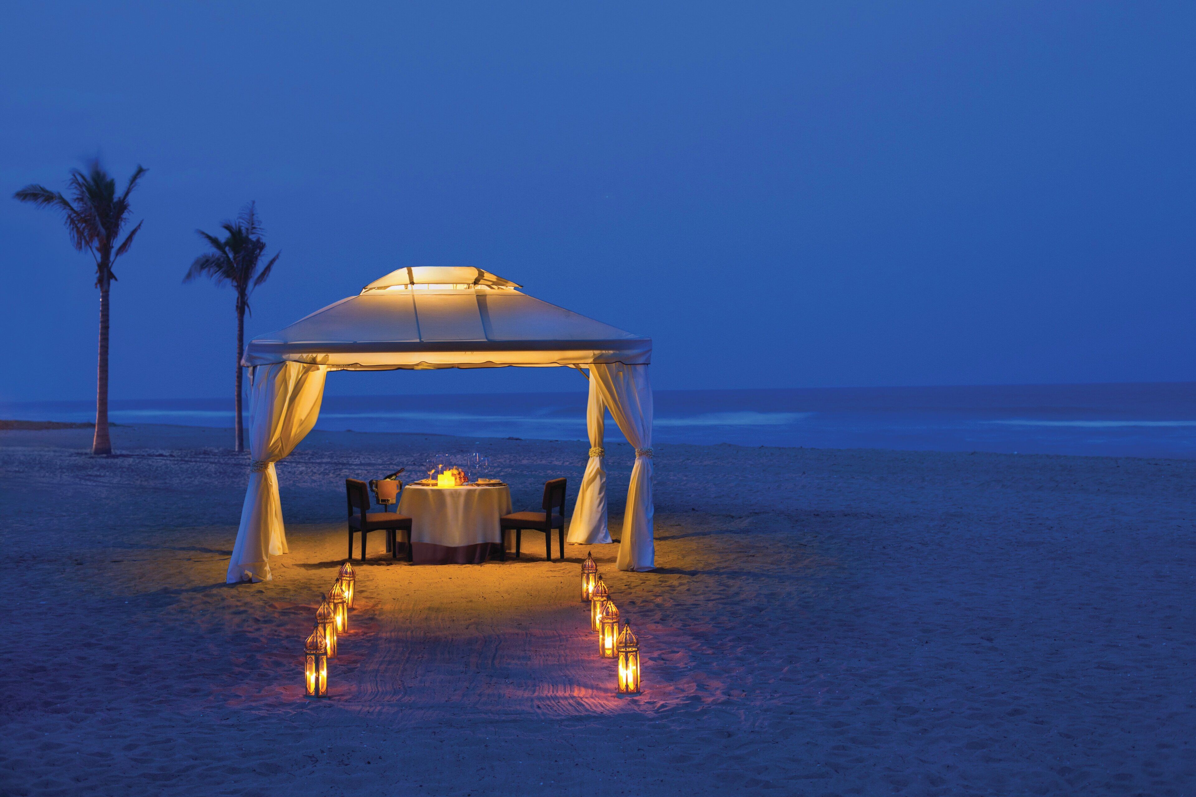 A beach setting with a tent, a table set for two, and a row of lanterns leading to the table.