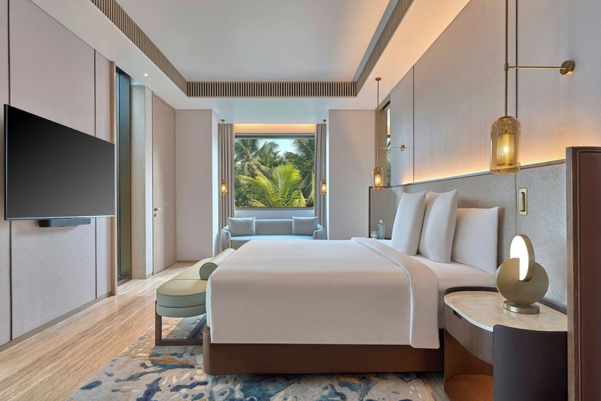 A modern hotel room with a large bed, a flat-screen TV, a seating area with a view of palm trees, and a large window.