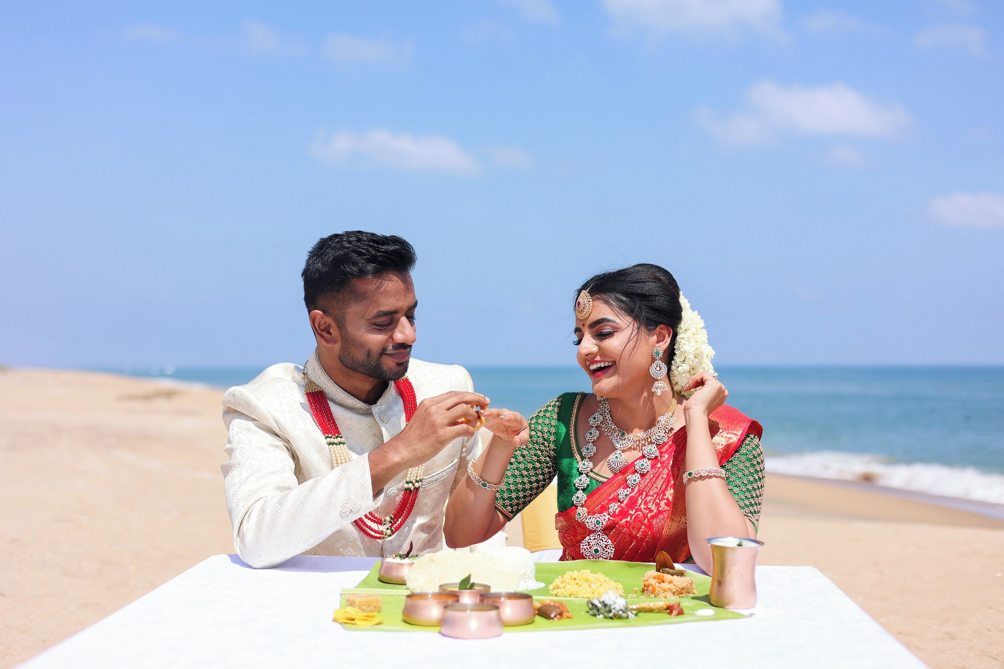 Ein Paar sitzt an einem Tisch mit traditionellem Essen am Strand, im Hintergrund das Meer.