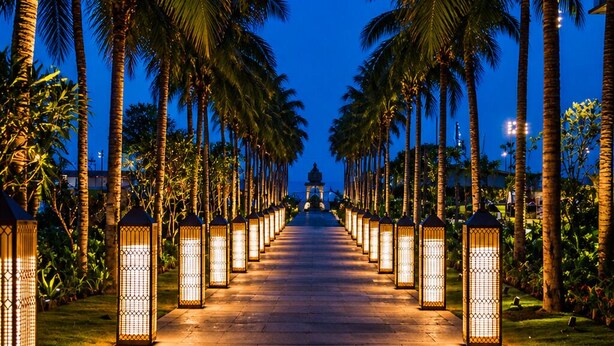 A pathway lined with palm trees and illuminated lanterns leading to a distant structure.