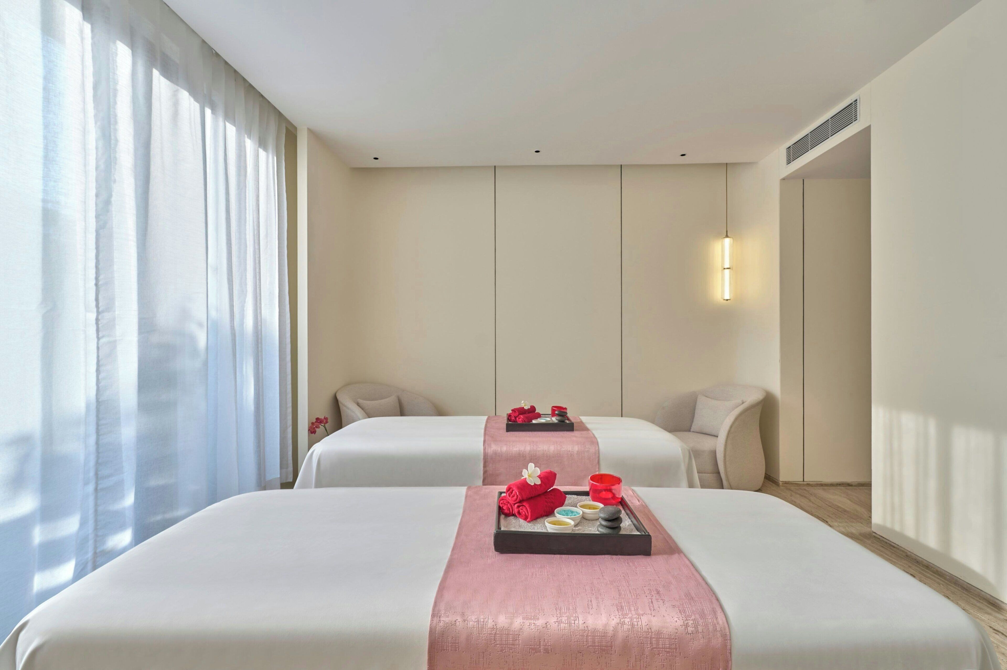 A hotel room with two beds, a large window with sheer curtains, a wall-mounted air conditioning unit, and a bedside table with a tray of amenities.